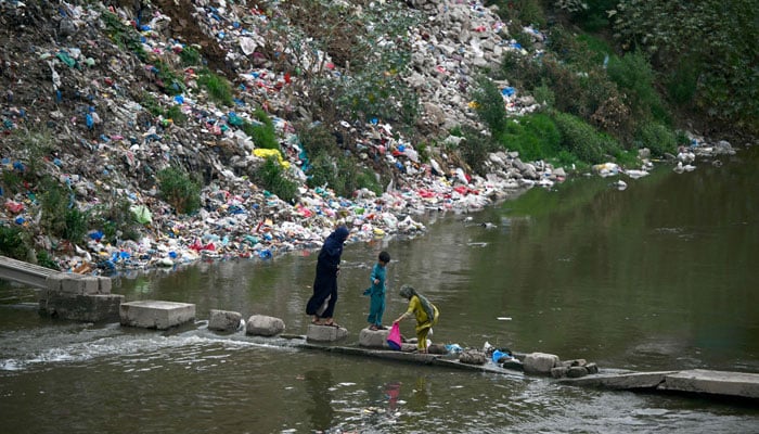 People cross a polluted stream running past a garbage dump on World Environment Day in Rawalpindi on June 5, 2024. — AFP