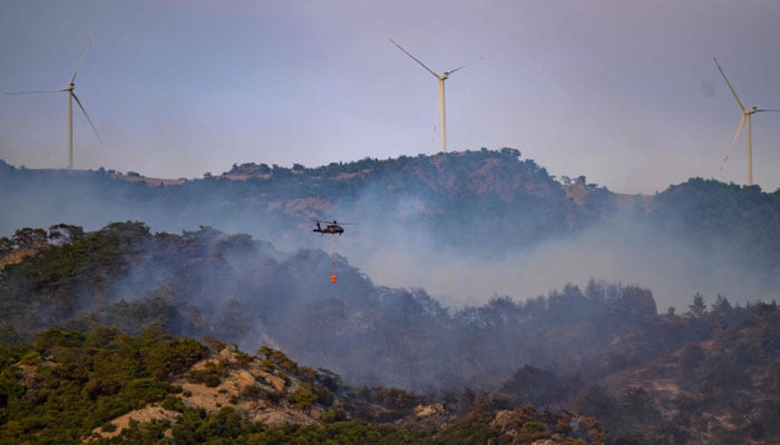 This photograph shows a helicopter carrying water to fight a forest fire, with windmills in the background in Turkeys western province of Izmir on August 17, 2024. — AFP