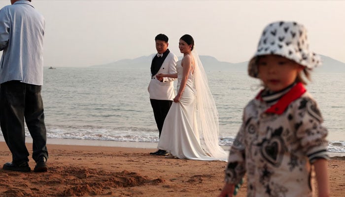 A child plays with sand near a couple taking part in a pre-wedding photoshoot on a beach in Qingdao, Shandong province, China April 21, 2024. — Reuters