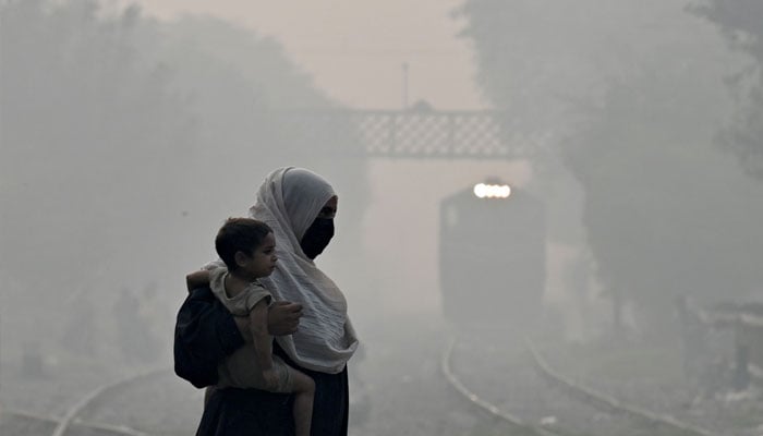 A woman carries a child as she walks across a railway track amid heavy smog in Lahore on October 29, 2024. — AFP