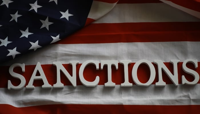 Representational image of sanctions written over a US flag. — Canva