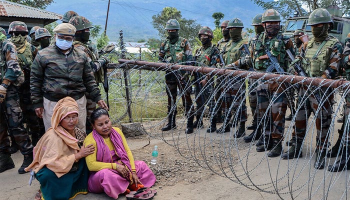 Family members sit in protest as army personnel stand guard at a checkpoint during a demonstration after a man from the Meitei community who worked at the camp allegedly remained missing in Imphal West on November 30, 2024. —AFP