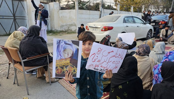 A child holding two placards in each hand is part of protest sit-in in progress in front of the Peshawar Press Club on Dec 27, 2024. — Facebook@Chinar