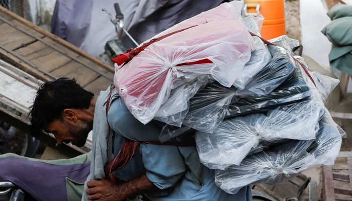 A labourer bends over as he carries packs of textile fabric on his back to deliver to a nearby shop in a market in Karachi on June 24, 2022. — Reyters