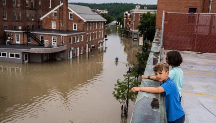 Two children observe the flooding from a building roof in downtown Montpelier, Vermont on Tuesday, July 11, 2023. One child is wearing a blue shirt and another is wearing a light blue shirt. Brown floodwaters surround brown brick buildings. Washington Post