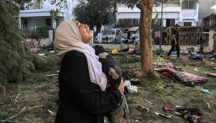 A woman, clutching a pillow, stands in the grounds of the al-Ahli Arab Hospital in Gaza City on 18 October 2023, surrounded by debris and personal effects from the previous nights massacre. — AFP