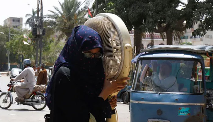 A woman carries a pedestal fan for repair during a hot and humid weather in Karachi. — Reuters