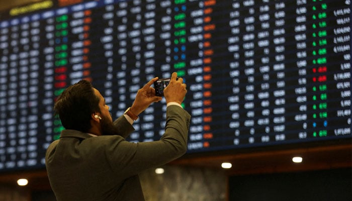 A man uses a mobile phone as he takes a photo of the electronic board displaying share prices during a trading session at the Pakistan Stock Exchange, on November 28, 2023. — Reuters