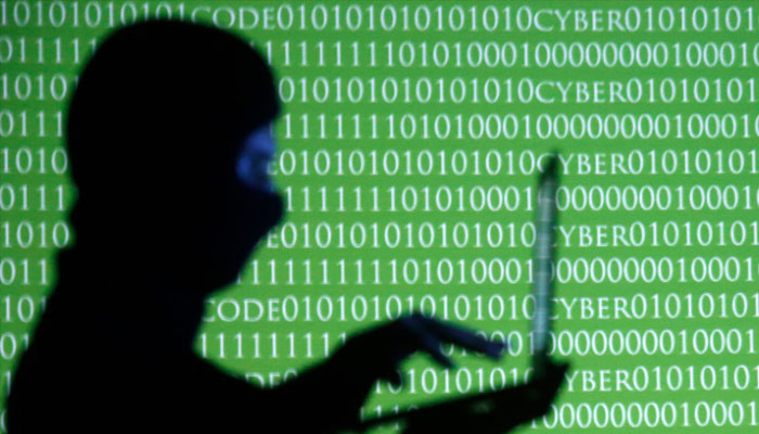 A person wearing a balaclava is silhouetted as he poses with a laptop in front of a screen projected with the word cyber and binary code, in this picture illustration taken in Zenica October 29, 2014. — Reuters