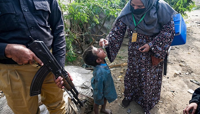 A police personnel stands guard as a a health worker administers polio drops to a child during a door-to-door poliovirus vaccination campaign at a slum area in Karachi on December 17, 2024. — AFP