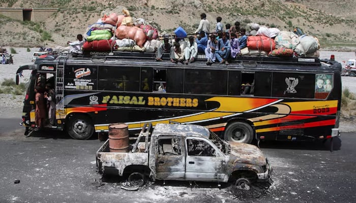 A bus with passengers sitting on the roof with belongings, drives past a damaged vehicle, following terrorist attacks in Bolan district of Balochistan, August 27, 2024. — Reuters